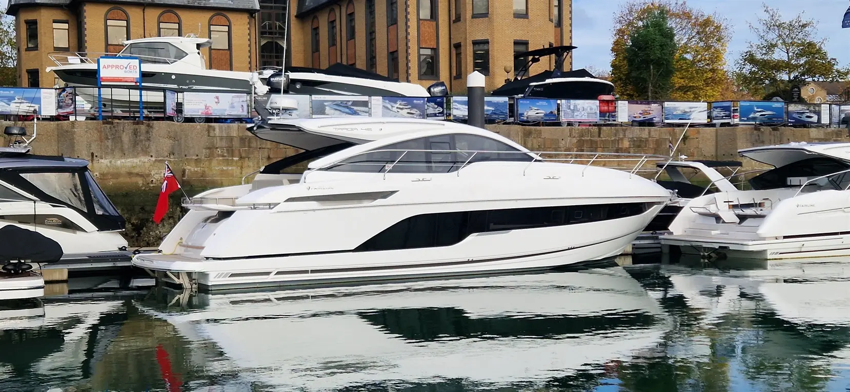 A sleek white motorboat is docked beside other vessels at a marina, reflecting in the calm water. "Approved Boats" sign visible.